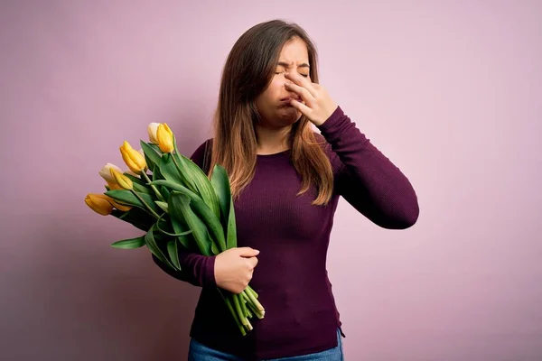 年轻的金发女人捧着一束浪漫的黄色郁金香花 背景是粉色的 身上散发着一股难闻 难以忍受的气味 她用手指捂住鼻子屏住呼吸 — 图库照片