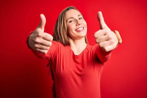 手で積極的なジェスチャーを行うことを承認孤立した赤い背景の上に立ってカジュアルなTシャツを着て若い美しいブロンドの女性 成功のために笑顔と幸せを親指アップ 勝者のジェスチャー — ストック写真