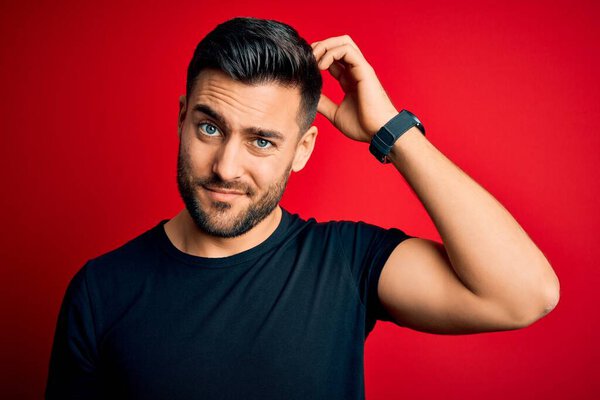 Young handsome man wearing casual black t-shirt standing over isolated red background confuse and wonder about question. Uncertain with doubt, thinking with hand on head. Pensive concept.