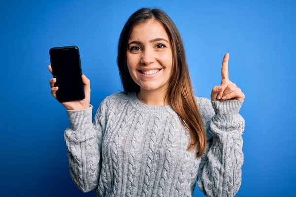 青い孤立した背景の上に空白のスマートフォンの画面を示す若い女性は 幸せな顔をしたアイデアや質問指差し指で驚いて ナンバーワン — ストック写真