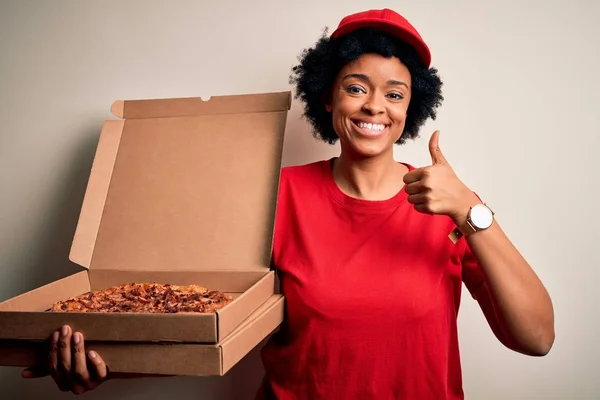 Young African American afro delivery woman with curly hair holding box with Italian pizza happy with big smile doing ok sign, thumb up with fingers, excellent sign