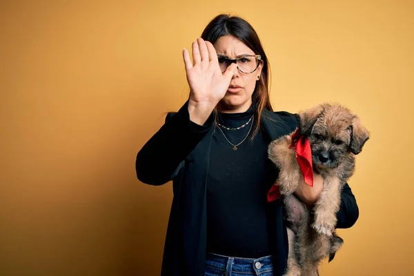 Young beautiful brunette woman holding cute puppy pet over isolated yellow background with open hand doing stop sign with serious and confident expression, defense gesture