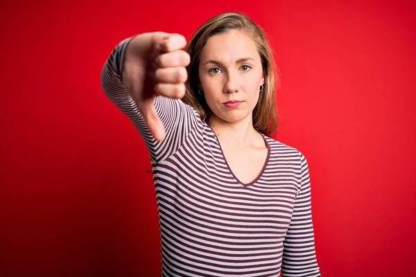 孤立した赤い背景にカジュアルなストライプのTシャツを着た若い美しいブロンドの女性は 親指ダウンジェスチャーで不満と怒りの拒絶と否定を示しています 悪い表現 — ストック写真