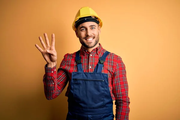 年轻的建筑工人身穿建筑制服 头戴安全帽 头戴黄色的隔离背景 带着四号手指 面带微笑 自信而快乐 — 图库照片