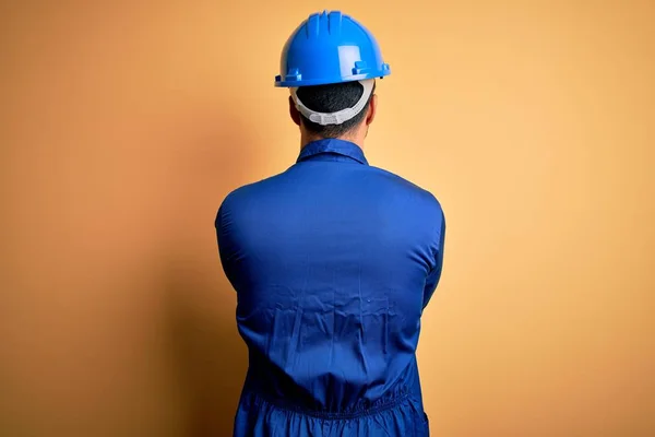 Mechanic man with beard wearing blue uniform and safety helmet over yellow background standing backwards looking away with crossed arms