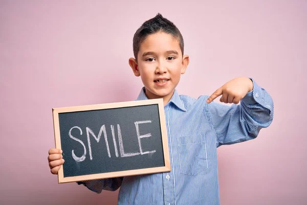 Young little boy kid showing blackboard with smile word as happy message over pink background with surprise face pointing finger to himself