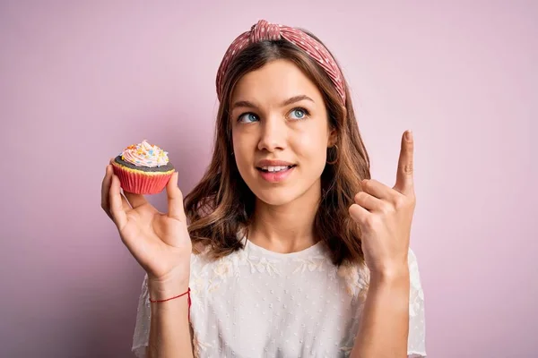 若いですブロンド女の子食べます甘いですチョコレートカップケーキ以上ピンク隔離された背景驚きでAアイデアや質問指差し指とともに幸せな顔 ナンバーワン — ストック写真