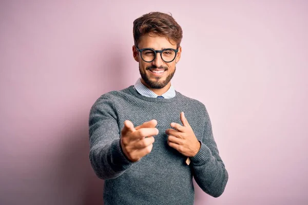 ひげを生やした若いハンサムな男は 幸せと面白い顔をしてカメラに指を指してピンクの背景の上に立って眼鏡とセーターを着ていた 良いエネルギーと振動 — ストック写真
