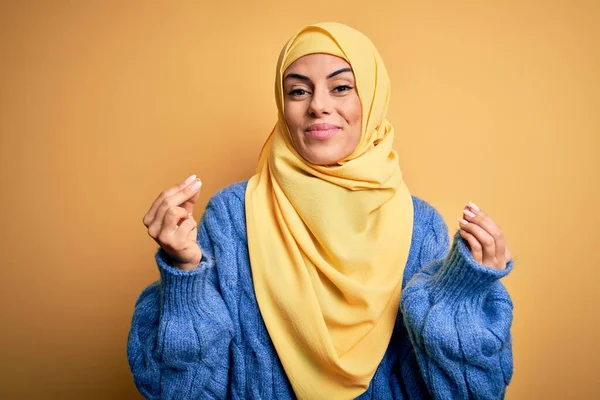 若いです美しいですブルネットイスラム教徒の女性身に着けていますアラブヒジャーブ上の隔離された黄色の背景に手でお金のジェスチャーを行います 給与支払いを求めます 億万長者ビジネス — ストック写真