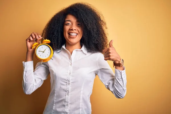 Young african american woman with afro hair holding classic alarm clock over yellow background happy with big smile doing ok sign, thumb up with fingers, excellent sign