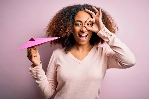 若いですアフリカ系アメリカ人女性とともにAfro毛保持紙飛行機上のピンクの背景とともに幸せな顔笑顔Doing Signとともに手で目を通して指を見て — ストック写真