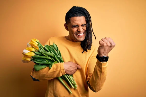 Young african american afro romantic man with dreadlocks holding bouquet of yellow tulips very happy and excited doing winner gesture with arms raised, smiling and screaming for success. Celebration concept. — Stock Image Young African American Afro Romantic Man Dreadlocks Holding Bouquet Yellow Royalty Free Stock Images