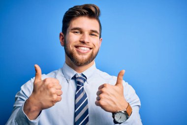 Sarışın, sakallı, mavi gözlü, şık gömlekli ve kravatlı genç bir iş adamı. Ayakta başarı işareti yapıyor. Elleriyle olumlu bir hareket yapıyor. Neşeli ifade ve kazanan jest.