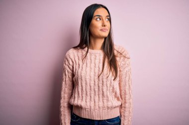 Genç, güzel, esmer bir kadın, pembe arka planda günlük süveter giyiyor. Gülümsüyor ve uzaklara bakıyor..