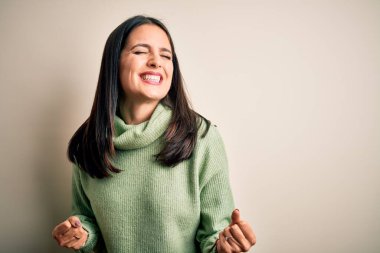 Mavi gözlü, balıkçı yaka kazak giyen genç esmer kadın çok mutlu ve heyecanlı bir şekilde kollarını kaldırmış, gülümseyen ve başarı için çığlık atan hareketler yapıyor. Kutlama konsepti.