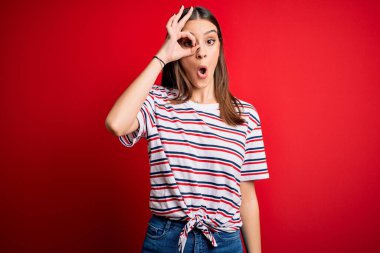 Genç, güzel, esmer, sıradan çizgili tişört giyen, kırmızı arka planda duran, şaşırmış yüzüyle şaşırmış, parmaklarından bakan bir jest yapan bir kadın. İnançsız ifade.