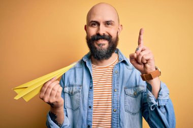 Sarı arka plan üzerinde kağıt uçak tutan sakallı yakışıklı kel adam bir fikre ya da soru işaretine şaşırdı.
