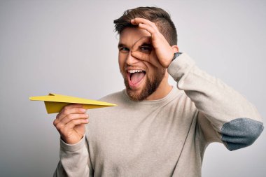 Sarışın, sakallı, mavi gözlü, beyaz arka planda kağıt uçak tutan, gülümseyen bir genç adam.