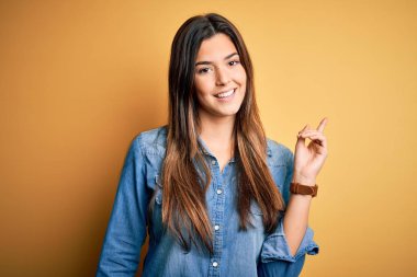 Genç, güzel bir kız, sıradan bir kot gömlek giyiyor, sarı arka planda duruyor, yüzünde kocaman bir gülümseme, eli ve parmağı kameraya bakıyor..