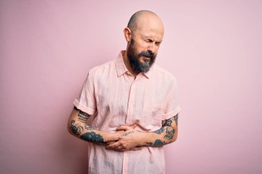 Sakallı ve dövmeli yakışıklı kel bir adam. İzole edilmiş pembe arka planda günlük gömlek giyiyor. Midesi bulanıyor, acı verici bir hastalığı var. Ache kavramı.