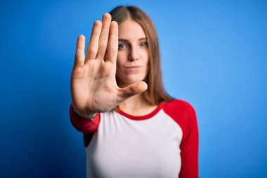 Young beautiful redhead woman wearing casual t-shirt over isolated blue background doing stop sing with palm of the hand. Warning expression with negative and serious gesture on the face. clipart