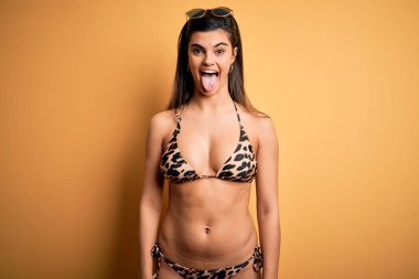 Genç ve güzel esmer kadın tatilde. Sarı arka planda mayo bikini giyiyor. Dilini komik bir ifadeyle dışarı çıkarıyor. Duygu kavramı.