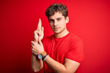 Sarışın, kıvırcık saçlı, kırmızı arka planda sıradan tişörtler giyen, el hareketleriyle sembolik silah tutan, cinayet silahlarıyla oynayan, sinirli yüzlü genç bir adam.