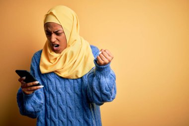Genç Afrikalı Afrikalı Afro kadın, başörtüsü takıyor. Akıllı telefon kullanıyor. Sinirli ve sinirli bir şekilde bağırıyor.