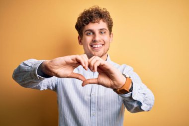 Sarışın, kıvırcık saçlı, sarı arka planda çizgili tişört giyen, kalp sembolü ve elleri ile şekil veren genç, sarışın bir adam. Romantik konsept.