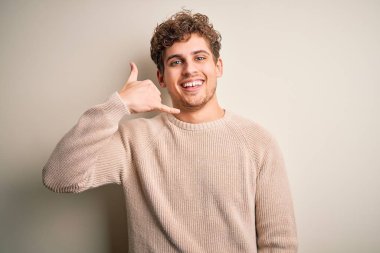 Sarışın, kıvırcık saçlı, beyaz arka planda rahat süveter giyen, telefonla el ve parmaklarıyla telefonla konuşan genç, yakışıklı bir adam. İletişim kavramları.