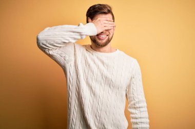 Sarışın, sakallı ve mavi gözlü genç adam sarı arka planda beyaz kazak giyiyor gözleri elle örtüyor, ciddi ve üzgün görünüyor. Görme, saklama ve reddetme konsepti