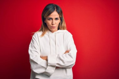 Orta yaşlı güzel sporcu kadın izole edilmiş kırmızı arka plan şüphecisi ve gergin, kollarını çapraz yapıp onaylamayan bir ifade takınıyor. Negatif kişi.