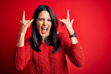 Mavi gözlü genç esmer kadın izole edilmiş kırmızı arka planda günlük süveter giyiyor çılgın bir ifadeyle elleri havada rock sembolü yapıyor. Müzik yıldızı. Ağır müzik konsepti.