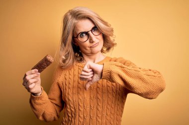 Orta yaşlı güzel sarışın kadın sarı arka planda enerjik protein çubuğu yiyor. Kızgın yüzü var. Eksi işareti var.