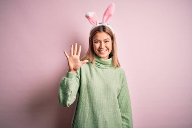 Paskalya tavşanı kulakları takan genç ve güzel bir kadın pembe arka plan üzerinde duruyor ve beş numaralı parmaklarıyla işaret ederken kendinden emin ve mutlu bir şekilde gülümsüyor..