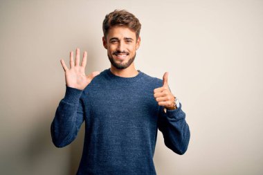Sakallı, sıradan bir süveter giyen, beyaz arka plan üzerinde duran, altı numaralı parmaklarıyla işaret eden, kendinden emin ve mutlu gülümseyen genç yakışıklı bir adam..