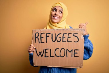 İslami tesettüre bürünmüş güzel bir Arap kadın elinde mültecilerle bayrak tutuyor. Hoş geldin mesajı gösteriyor ve gülümseyen bir gülümsemeyle baş parmağıyla işaret ediyor.