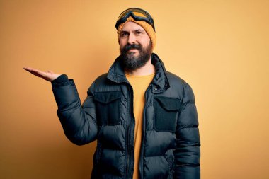 Sakallı, kar maskeli, kar maskeli ve kayak gözlüklü yakışıklı kayakçı. Neşeli bir şekilde sunum yapıyor ve avuç içi kameraya bakıyor..