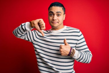 İzole edilmiş kırmızı arka planda dikilen sıradan çizgili tişört giyen genç Brezilyalı bir adam. Baş parmaklarını yukarı aşağı hareket ettiriyor, anlaşmazlık ve anlaşma ifadesi takınıyor. Çılgın çatışma