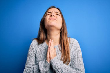 Güzel, genç bir kadın, mavi izole edilmiş arka planda dikilmiş yün kazak giyiyor. Elleriyle yalvarıyor ve dua ediyor. Yüzünde umut ifadesi var. Çok duygusal ve endişeli. Yalvarıyorum..