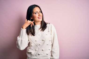 Mavi gözlü genç esmer kadın izole edilmiş pembe arka planda rahat bir süveter giyiyor telefonla el ve parmaklarıyla telefonla konuşuyor gibi gülümsüyor. İletişim kavramları.