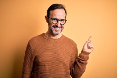 Orta yaşlı, beyaz, kahverengi süveter giyen ve gözlüklü, izole edilmiş sarı arka planda, yüzünde kocaman bir gülümseme olan, elini kameraya doğru gösteren bir adam..