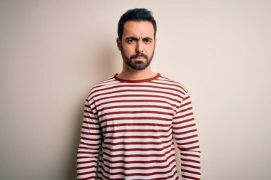 Sakallı, sıradan çizgili tişörtlü, beyaz arka planda duran, kuşkucu ve sinirli, sorun yüzünden üzgün olan genç yakışıklı bir adam. Negatif kişi.