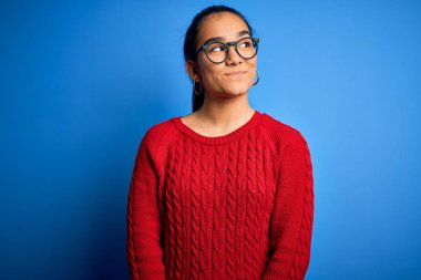 Genç, güzel Asyalı bir kadın mavi arka planda gözlük ve süveter takıyor gülümsüyor ve başka tarafa bakıyor..