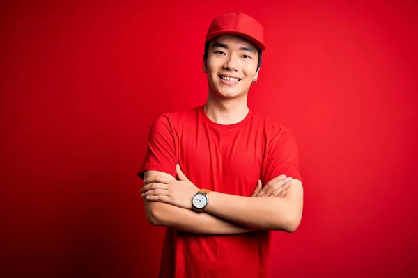 Young handsome chinese delivery man wearing cap standing over isolated red background happy face smiling with crossed arms looking at the camera. Positive person.