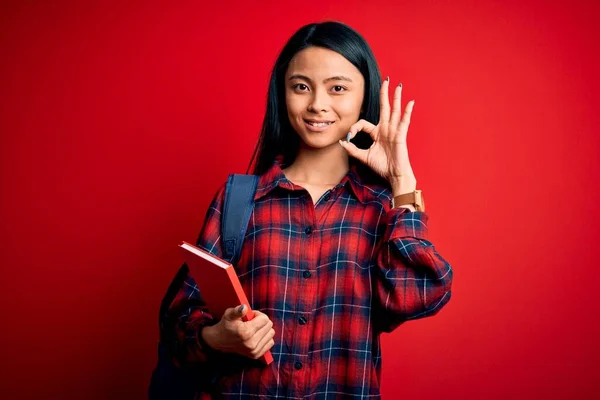 Young beautiful chinese student woman holding book standing over isolated pink background doing ok sign with fingers, excellent symbol