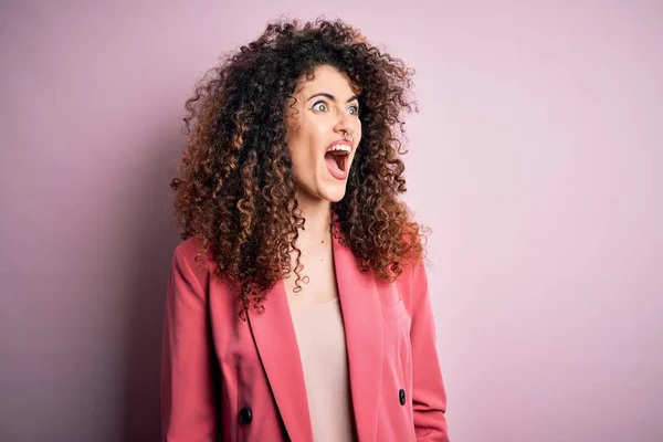 Young beautiful businesswoman with curly hair and piercing wearing elegant jacket angry and mad screaming frustrated and furious, shouting with anger. Rage and aggressive concept.