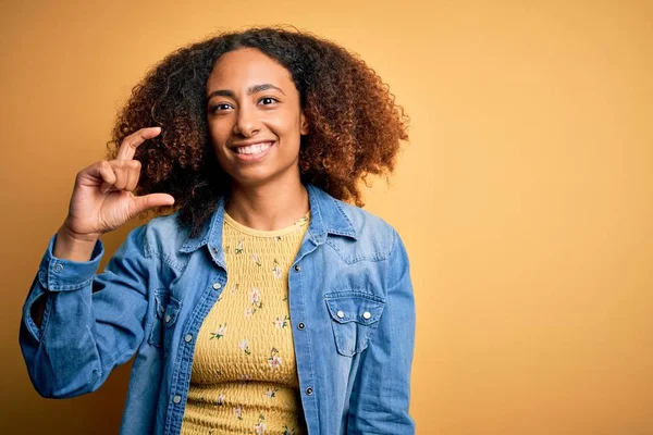 黄色の背景にカジュアルなデニムシャツを着てアフロの髪を持つ若いアフリカ系アメリカ人女性は笑顔と指を見てとカメラと小さなサイズの看板を行う手で自信を持ってジェスチャー 概念の測定 — ストック写真