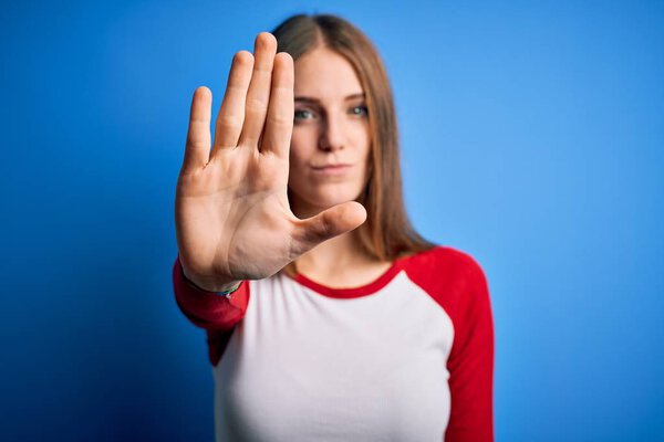 Young beautiful redhead woman wearing casual t-shirt over isolated blue background doing stop sing with palm of the hand. Warning expression with negative and serious gesture on the face.