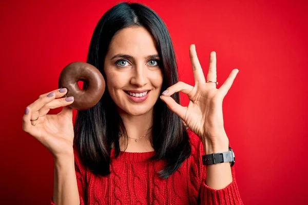 年轻的黑发女人 蓝眼睛 吃巧克力甜甜圈 背景是红色的 背景是孤立的 手握手签 象征非凡 — 图库照片